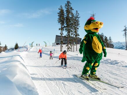 Dragon Park: Schmidolin skiing alongside children in the snow. | © Schmittenhöhebahn AG/Punkt und Komma