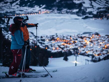 Zwei Skifahrer auf einem Hang, mit beleuchtetem Dorf und See im Hintergrund. | © Schmittenhöhebahn AG/Zooom-Productions