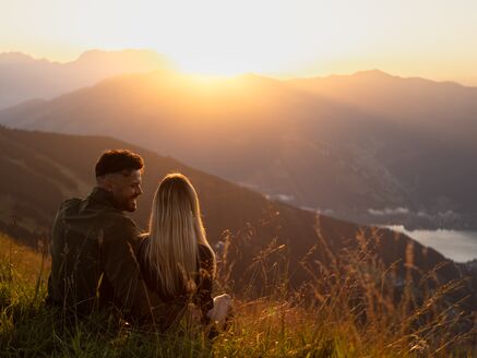 Paar sitzt im Gras bei Sonnenaufgang mit Blick Richtung Sonne | © Kuscheiart